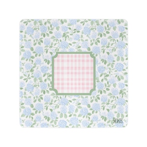 May include: A square decorative plate featuring a repeating pattern of light blue hydrangea flowers and green leaves. In the center, a square panel with a pink and white gingham check pattern is framed by a sage green border. The letters MWH are printed.