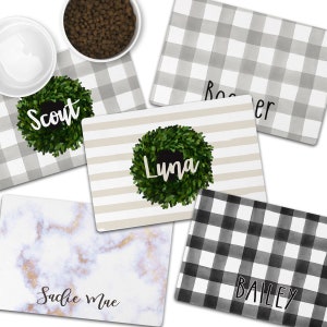 May include: Five different pet food mats with various designs. The mats are white with black and gray checkered patterns, white with gray stripes, white with a marble pattern, and white with a green wreath. The mats have the names "Scout", "Luna", "Bailey", "Rooster", and "Sadie Mae" written on them.