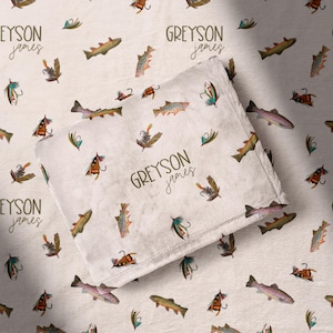 May include: A light beige blanket with a repeating pattern of watercolor fish and fishing lures. The blanket has the name "GREYSON james" printed on it in a cursive font. The background also has the same pattern.