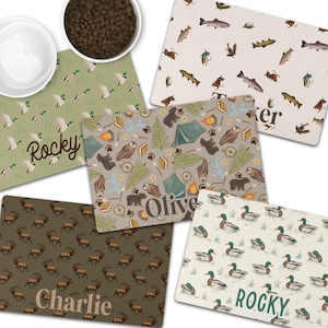 May include: A collection of personalized pet food mats with various nature-themed patterns. Designs include ducks, fish, elk, and camping scenes, each with a pet's name. The mats are rectangular and feature a neutral color palette.