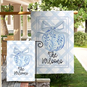 May include: Two blue and white garden flags with a watercolour design. Each flag features a blue bow and a floral ornament with the text "The Wilsons" in a script font. The background is a light blue and white gingham pattern.