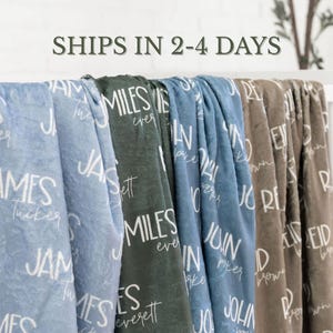 May include: A collection of soft, plush blankets in various colors including light blue, dark green, and brown. Each blanket features a repeating pattern of personalized names printed in a white, handwritten-style font across the fabric surface.