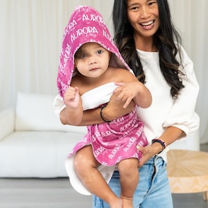 May include: A hooded towel in a vibrant magenta color featuring a repeating white script pattern of the name Aurora. The towel has a white interior lining and is designed to wrap around the body with a hood to cover the head area.