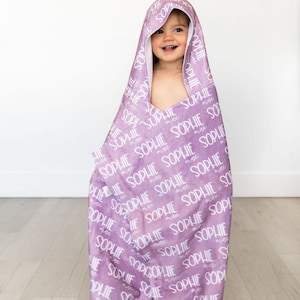 May include: A hooded baby towel in a soft lavender hue, featuring the name "Sophie" in a repeating white script. The towel has a hood and is designed to wrap around a child. The background is a plain white wall and light-colored wood floor.