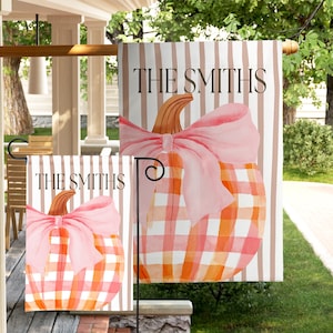 May include: Two garden flags featuring a watercolor pumpkin with a pink bow and a buffalo check pattern in orange and white. The background has vertical brown stripes, and the text THE SMITHS is printed in black at the top of each flag.