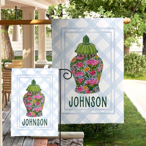 May include: Two garden flags with a floral vase design. The vase is adorned with pink and yellow flowers and a green lid, set against a light blue plaid background. The name "JOHNSON" is printed in green below the vase.