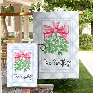 May include: A decorative garden flag featuring a watercolor design of a red bow and mistletoe on a light blue plaid background. The flag includes the text "The Smiths" in a script font. The flag is displayed outdoors.