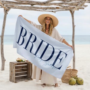 May include: A light blue beach towel with the word "BRIDE" in navy blue block letters. Below the word "BRIDE" is the text "Mrs. Johnson" in a lighter blue script. The towel is held up on a beach.