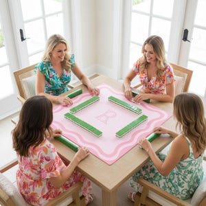 May include: A square mahjong mat in light pink with a wavy white border and a central letter B, featuring green and white mahjong tiles arranged in a square formation on the surface, set upon a wooden table with four chairs around the perimeter.