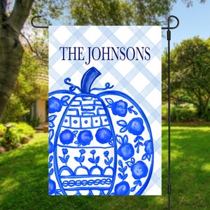 May include: A rectangular garden flag featuring a light blue and white plaid background with the text THE JOHNSONS in dark blue. A large, central pumpkin illustration is decorated with blue floral patterns and geometric lines on a white base.