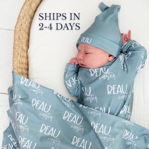 May include: A newborn baby wearing a light blue hat and matching sleepsuit, with the name "Beau Carter" printed on the outfit and blanket. The baby is lying on a white surface inside a woven basket.