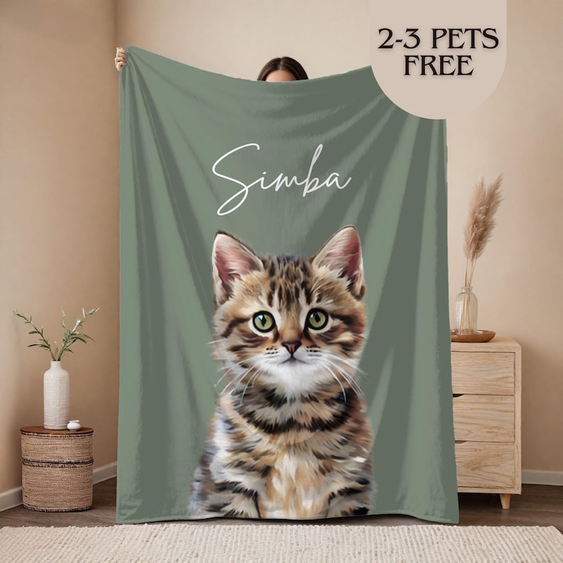 May include: A green blanket with a detailed illustration of a tabby kitten. The kitten's name, "Simba," is written in cursive. The blanket also has the text "2-3 PETS FREE" in a circular design.