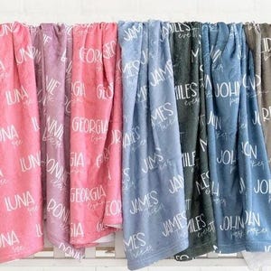 May include: A collection of soft blankets in various colors including pink, lavender, blue, dark gray, and brown. Each blanket features a repeating pattern of white printed names such as Luna, Bonnie, Georgia, James, Miles, and John.