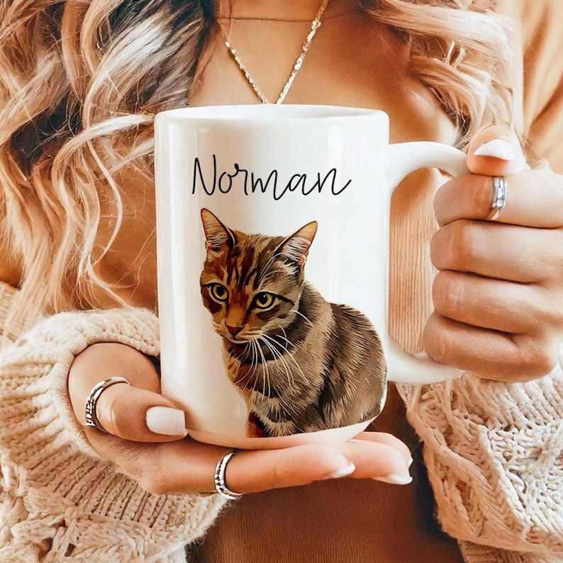 May include: White ceramic mug featuring a detailed illustration of a tabby cat with brown and tan stripes. The name "Norman" is written above the cat in black script. The mug has a large handle.