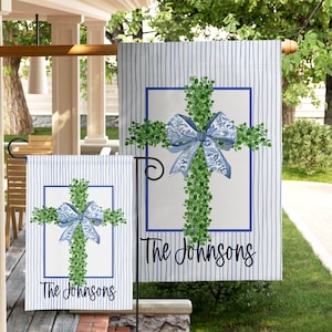 May include: Two outdoor garden flags with a cross design. The cross is made of green foliage and has a blue bow in the center. The flags have a blue and white striped background and the text "The Johnsons".