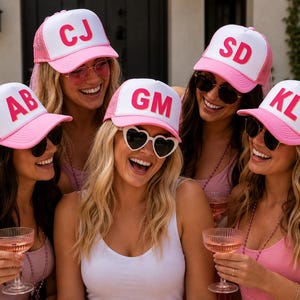 May include: Five pink and white trucker hats featuring mesh backs and attached pink veils. Each hat displays two large, bold, hot pink capital letters on the front white panel, specifically the combinations AB, CJ, GM, SD, and KL, in a sans-serif font.
