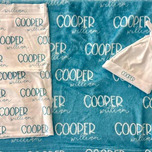 K&ouml;nnte beinhalten: Eine wei&szlig;e Puckdecke und eine passende wei&szlig;e Babym&uuml;tze, beide mit den in T&uuml;rkis gedruckten Namen Cooper und William. Diese Artikel liegen auf einer t&uuml;rkisfarbenen Decke, die ebenfalls die Namen Cooper und William in wei&szlig;em Text zeigt.