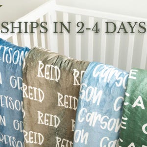 May include: Four soft blankets draped over a white crib. The blankets are blue, brown, light blue, and green, each featuring a repeating white name printed in a bold, sans-serif font. The names displayed are Greyson, Reid, Carson, and Cade.