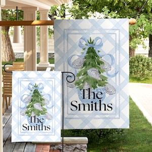 May include: A garden flag and a smaller flag feature a watercolor design of a Christmas tree decorated with oyster shells and a blue bow. The flags have a light blue plaid background and the name "The Smiths" in black.