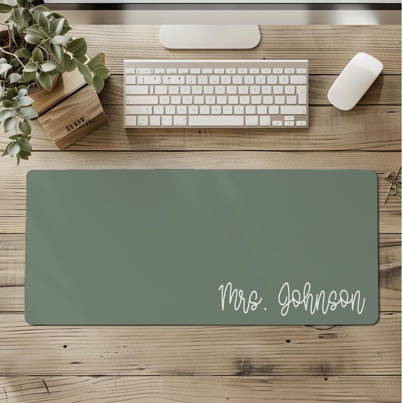Puede incluir: Alfombrilla de escritorio verde salvia rectangular con el texto "Mrs. Johnson" en blanco. La alfombrilla est&aacute; sobre un escritorio de madera, junto con un teclado, un rat&oacute;n y una peque&ntilde;a planta. Accesorio de oficina pr&aacute;ctico.