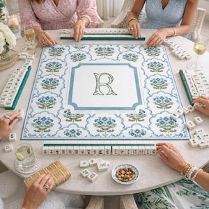 May include: A square mahjong game board featuring a white background with a repeating blue and green floral pattern, a central green letter R, and blue border text. The board is surrounded by white mahjong tiles with green backs and game pieces.