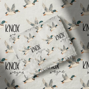 May include: A light gray baby blanket with a repeating pattern of flying ducks. The blanket is personalized with the name "KNOX" and the word "wayne" in a handwritten style font.