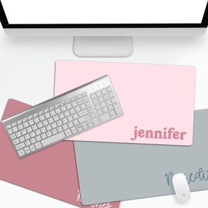 Puede incluir: Un alfombrilla de rat&oacute;n rosa claro personalizada con el nombre "Jennifer" en una fuente rosa a juego. Un teclado plateado y un rat&oacute;n blanco est&aacute;n sobre el escritorio. Se ven otras alfombrillas de rat&oacute;n de diferentes colores.