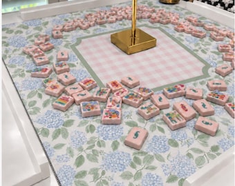 以下が含まれることがあります： 水色と緑のアジサイの花柄が描かれた正方形のゲームマットで、中央にはピンクと白のギンガムチェックの四角形があります。さまざまなカラフルなイラストやシンボルが描かれた、小さな長方形の薄いピンク色のタイルがマットの上に散らばっています。