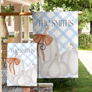 May include: A rectangular garden flag featuring a light blue and white plaid pattern, white pumpkins, and an orange bow. The text THE SMITHS is printed in black at the top. The design is displayed on a metal stand, showing the front and back view.