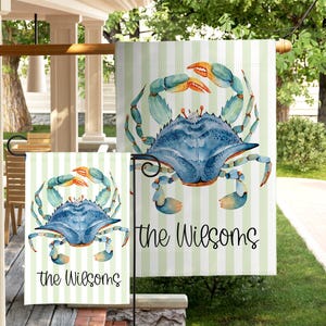 May include: Two garden flags with a watercolor crab design. The crab is blue with orange claws, set against a green and white striped background. The text "the Wilsoms" is printed in black script on both flags. The flags are displayed outdoors.