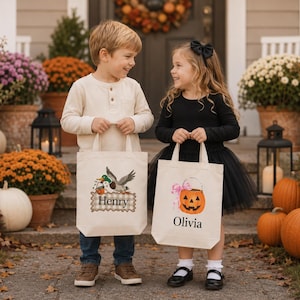 May include: Two off-white canvas tote bags featuring personalized names and Halloween graphics. One bag displays a mallard duck with a pumpkin bucket and the name Henry. The other bag features a jack-o'-lantern with a pink bow and the name Olivia.