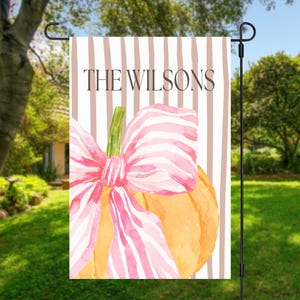 May include: A rectangular garden flag featuring a vertical brown and white striped background with the text THE WILSONS in brown. An orange pumpkin is centered at the bottom, adorned with a large, pink and white striped watercolor bow.