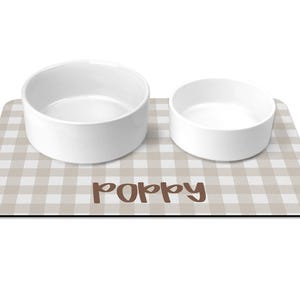 May include: A rectangular pet feeding mat with a beige and white gingham pattern featuring the name Poppy in brown script text. Two plain white ceramic bowls of different sizes are placed on top of the mat for food and water service.