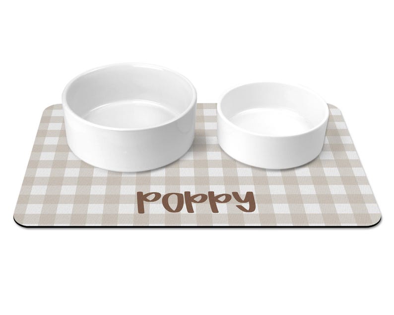 May include: A rectangular pet feeding mat with a beige and white gingham pattern featuring the name Poppy in brown script text. Two plain white ceramic bowls of different sizes are placed on top of the mat for food and water service.
