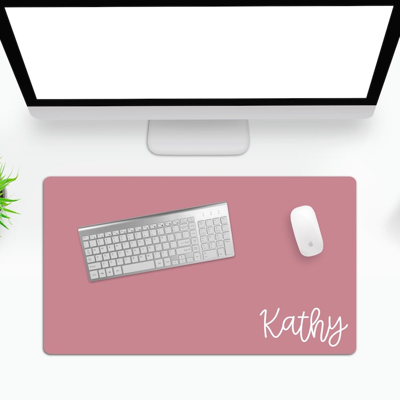 Puede incluir: Una alfombrilla de escritorio rosa polvoriento con un teclado plateado, un rat&oacute;n blanco y el nombre "Kathy" en escritura blanca. El teclado est&aacute; centrado en la alfombrilla, con el rat&oacute;n a la derecha. Un monitor de ordenador est&aacute; en el fondo.