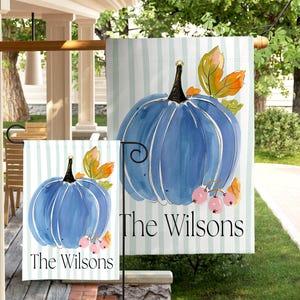 May include: Two garden flags featuring a watercolor blue pumpkin with a black stem, orange and green leaves, and pink berries against a light blue striped background. The text "The Wilsons" is printed in black at the bottom of each flag.