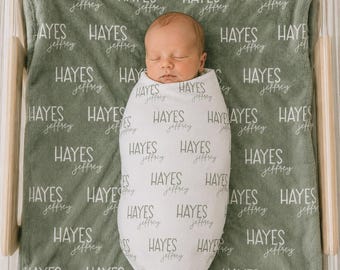 May include: A sage green baby blanket featuring a repeating pattern of the name Hayes Jeffrey in white text, with a swaddled infant centred on the blanket. The infant is wrapped in a white swaddle blanket also printed with the name Hayes Jeffrey.
