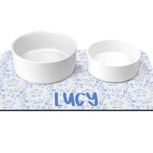 May include: White ceramic pet bowls with a blue floral design on a white background. The mat has the name "Lucy" printed in blue.