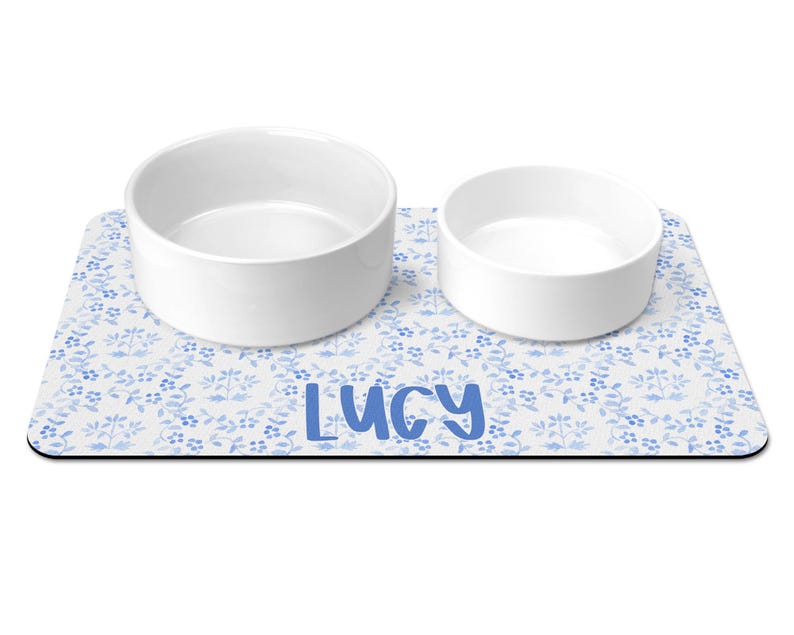 May include: White ceramic pet bowls with a blue floral design on a white background. The mat has the name "Lucy" printed in blue.