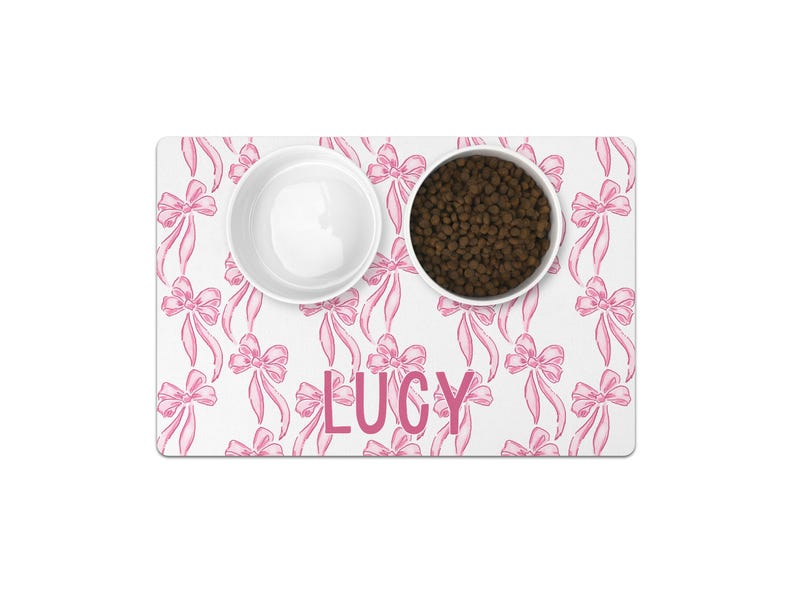 May include: White pet food mat with a pink bow pattern and the name "LUCY" printed in pink letters. The mat has two white bowls, one filled with brown pet food.