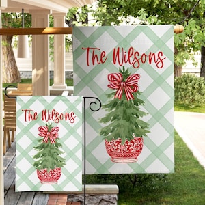 May include: Two outdoor Christmas garden flags. Each flag features a green Christmas tree with a red bow and pot, set against a green and white plaid background. The text "The Wilsons" is written in red script.