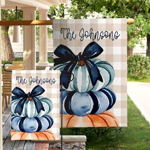 May include: A decorative garden flag featuring a stack of watercolor pumpkins in shades of blue, green, and orange, topped with a navy blue bow. The flag has the text "The Johnsons" in a script font.