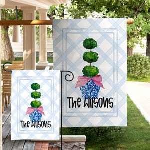 May include: A blue and white checkered garden flag with a topiary design. The topiary is green with a pink bow and sits in a blue floral pot. The text "The Wilsons" is printed in black.