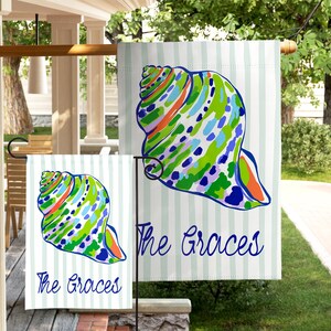 May include: Two rectangular garden flags featuring a light blue and white vertical striped background. Each flag displays a large, colorful seashell illustration in shades of green, blue, and orange, with the text The Graces in blue script.