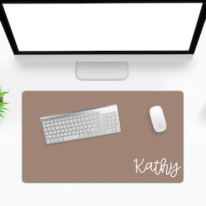 Puede incluir: Alfombrilla de escritorio rectangular de color topo con un teclado blanco, un rat&oacute;n blanco y el nombre "Kathy" en escritura cursiva. El teclado es plateado y tiene una distribuci&oacute;n est&aacute;ndar. El rat&oacute;n es de dise&ntilde;o minimalista.