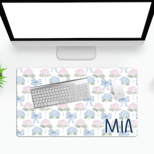 Puede incluir: Una alfombrilla de escritorio blanca con un patrón floral de hortensias azules y rosas y lazos azules. El texto "MIA" está impreso en azul en la esquina inferior derecha de la alfombrilla. Un teclado plateado y un ratón blanco están sobre la alfombrilla.