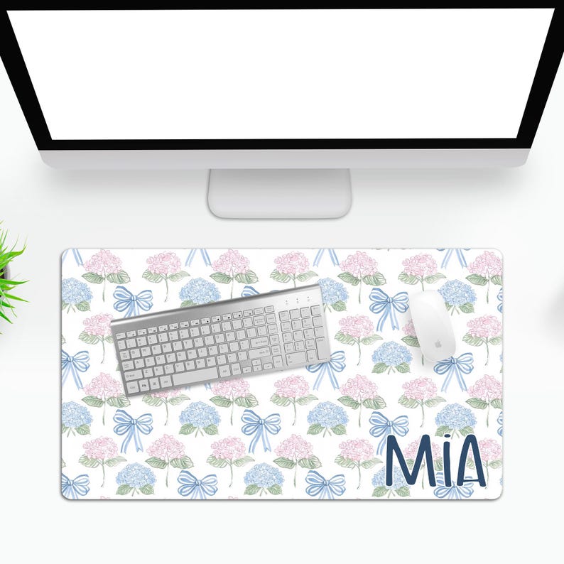 May include: A white desk mat with a floral pattern of blue and pink hydrangeas and blue bows. The text "MIA" is printed in blue at the bottom right corner. A silver keyboard and a white mouse are on the mat.