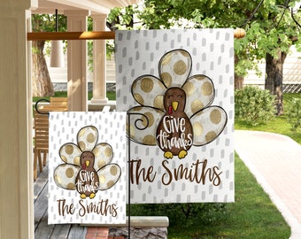 May include: Thanksgiving garden flag featuring a cartoon turkey with gold polka-dot feathers. The flag reads "Give thanks" and "The Smiths" in brown script against a white background with gray dashes. A smaller matching flag is also visible.