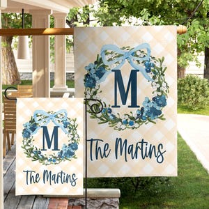 May include: Two garden flags with a light yellow and white checkered background. Each flag features a blue floral wreath with a blue bow and the letter "M". The text "The Martins" is below the wreath.