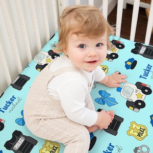 May include: A baby in a white long-sleeved shirt and beige overalls sits on a light blue crib sheet. The sheet features police-themed illustrations, including police cars, badges, handcuffs, and the name "Tucker Michael".
